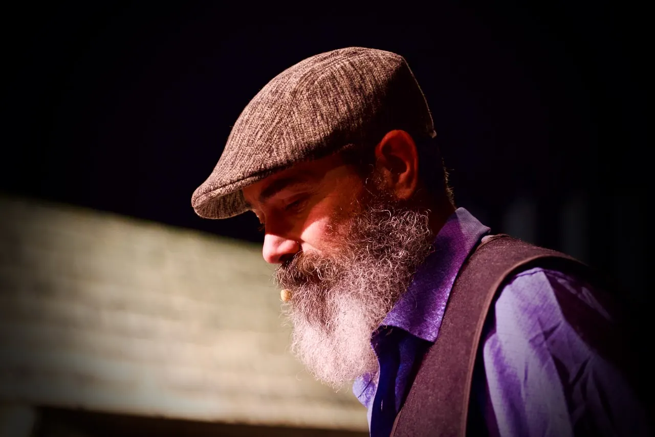 Tevye looks down in quiet thought, lit by warm stage light.