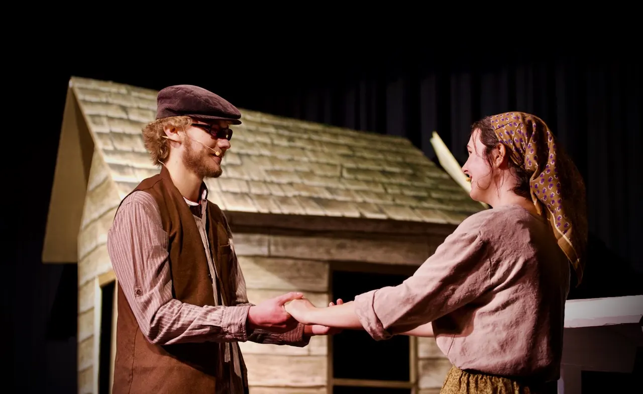 A young man and a young woman clasp hands in front of the cottage.
