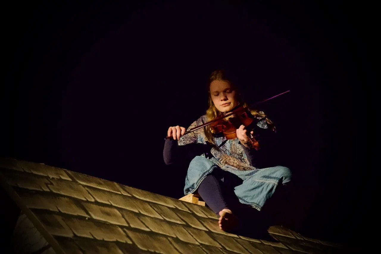 The fiddler, in a cap and patched clothes, sits on the peak of a cottage roof playing the violin under a single beam of light.