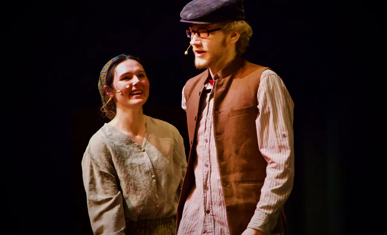 A laughing young woman stands beside a young man in glasses and a vest.