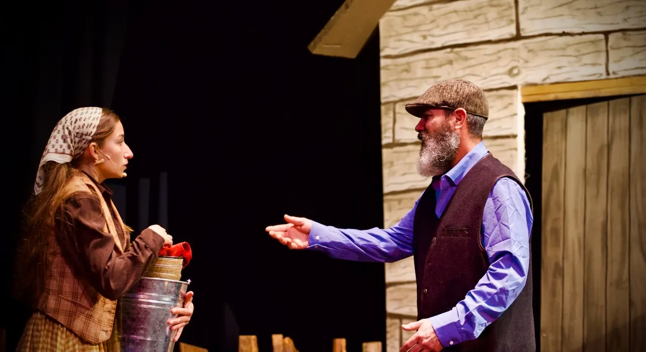 Outside the family cottage, Tevye gestures toward his daughter as she holds a milk can and flowers.